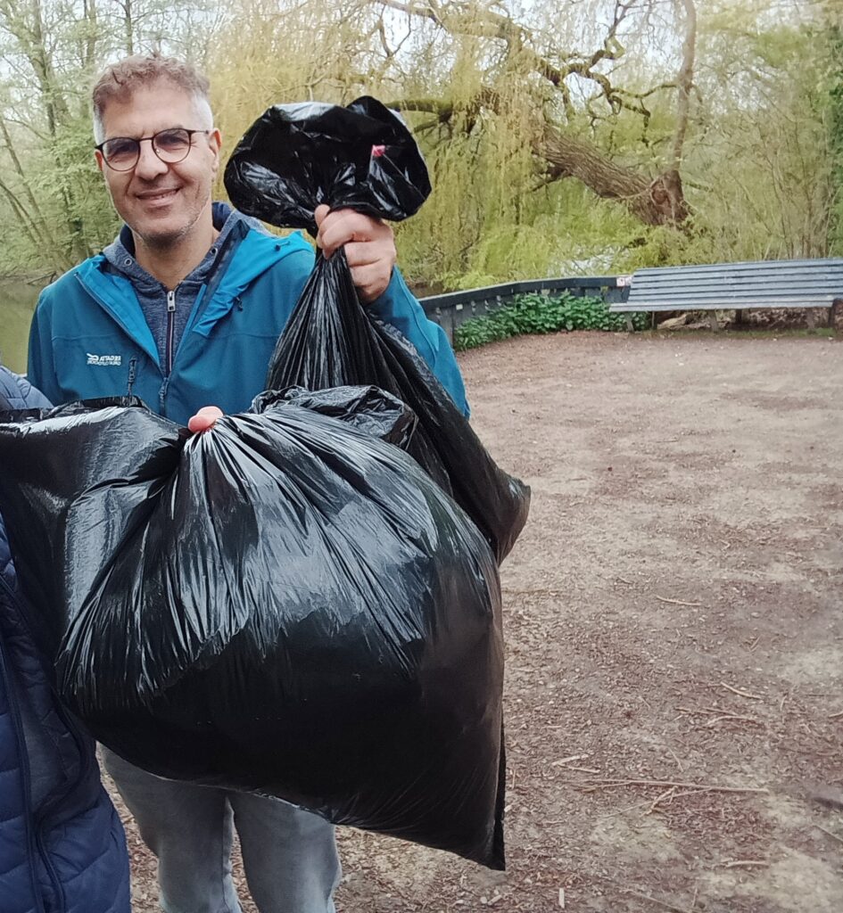 A man holds up a bag of trash with another person just in the foreground off screen holding up a second bag of trash they have collected from the ground.