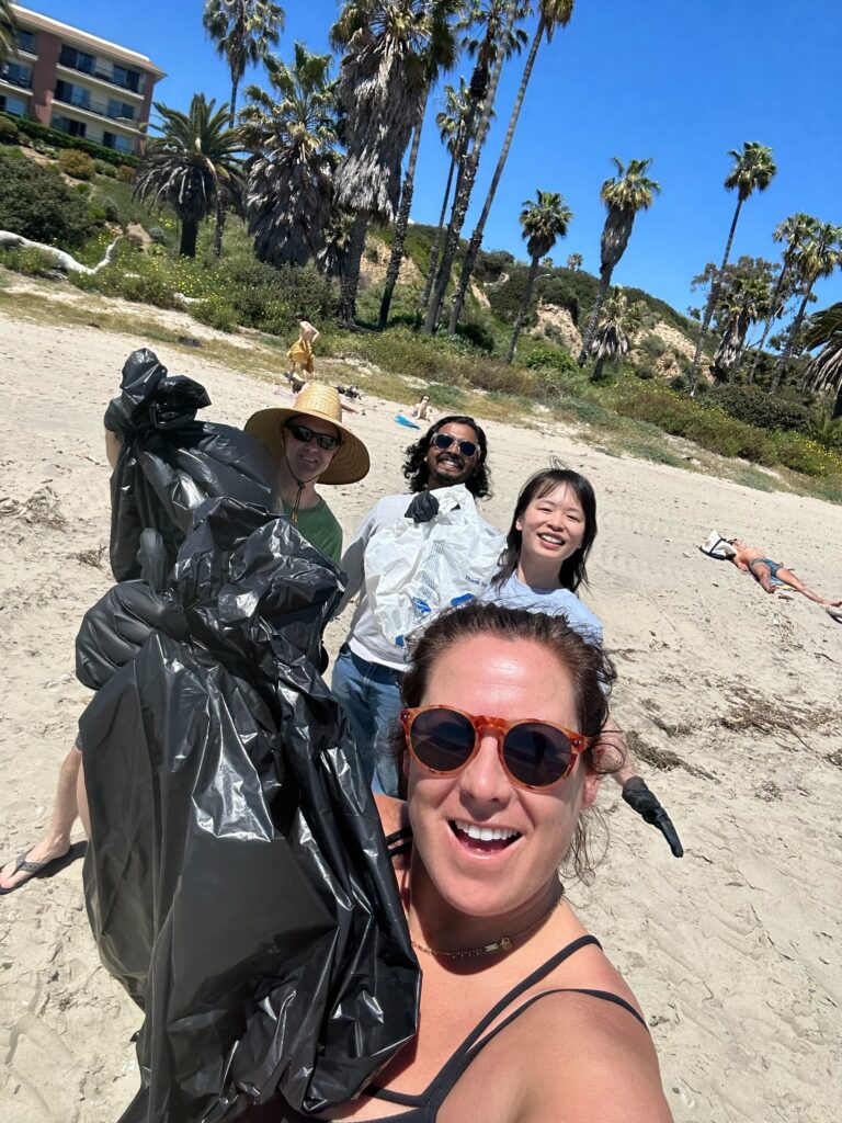 Four people on a beach hold up black trash bags and smile at the camera with palm trees behind them.